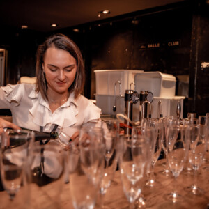 Photographie capturant l'élégance et l'énergie d'une festivité événementielle, avec des invités élégamment vêtus réunis dans un espace somptueusement décoré. Des lumières tamisées et des chandeliers suspendus créent une atmosphère intime et chaleureuse, tandis que des tables rondes drapées de nappes blanches sont parsemées de centres de table floraux délicats et de bougies scintillantes. Au fond, une scène accueille un groupe de musique, dont les mélodies douces et entraînantes invitent les convives à danser. Les rires et les conversations animées remplissent l'air, témoignant de moments de joie et de connexions partagées. Cette image illustre parfaitement le succès de l'événement, où chaque détail contribue à une expérience mémorable et raffinée pour tous les participants.